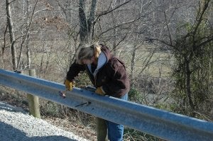 A female construction worker wearing heavy gloves uses a tool to repair or install a metal roadside guardrail next to a wooded area