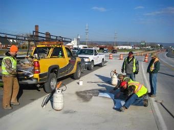 A road construction crew wearing high-visibility safety vests working on a pavement repair next to a yellow utility truck on a sunny day