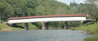 A side view of the historic Philippi Covered Bridge in West Virginia, featuring white wooden siding and a reddish-brown roof, spanning a wide river on stone piers against a lush, forested hillside