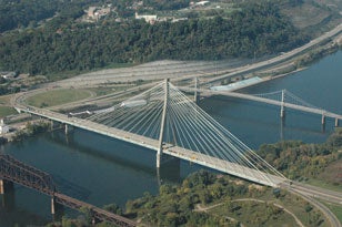 An aerial view of the Ohio River crossing at Weirton, West Virginia, featuring the single-tower, cable-stayed Veterans Memorial Bridge in the center, alongside a historic suspension bridge and a railway truss bridge