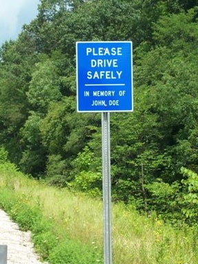 A blue roadside memorial sign on a metal post reading 'Please Drive Safely, In Memory of John Doe,' set against a background of lush green trees and roadside grass.