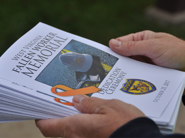 Hands holding a stack of printed programs for the West Virginia Fallen Worker Memorial Dedication Ceremony, with a small orange ribbon resting on top.