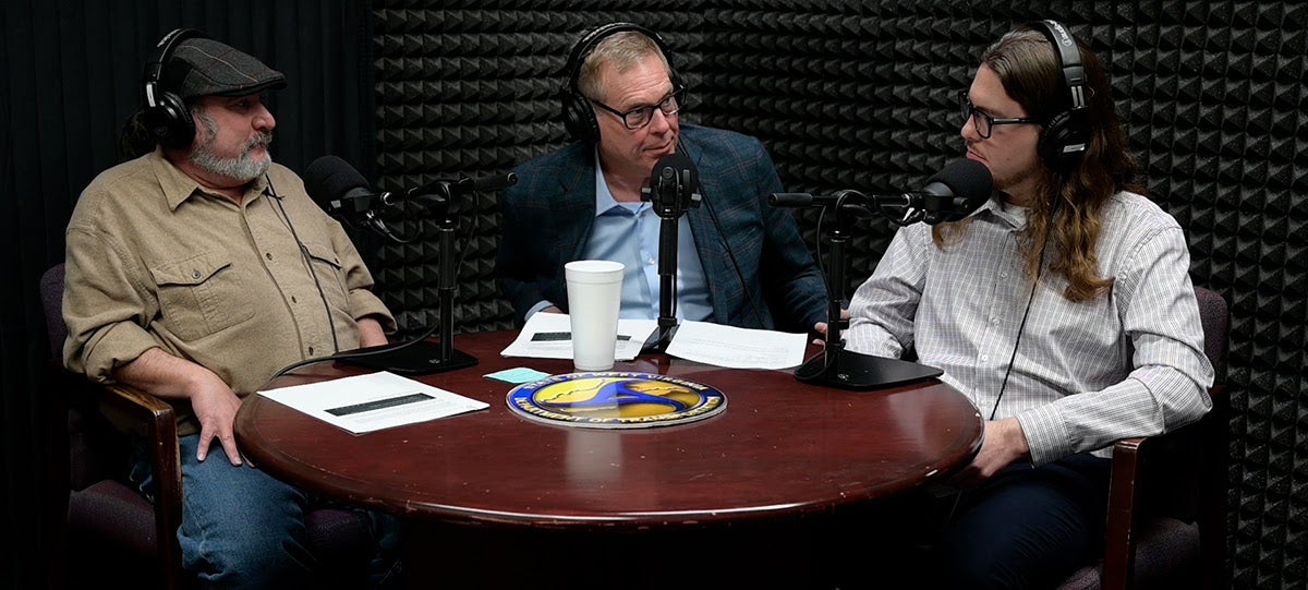 Three men wearing headphones sit around a table equipped with microphones, recording an episode of the WV on the DOT podcast in a soundproof studio. The table features the West Virginia Department of Transportation seal.