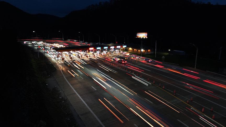 A high-angle, long-exposure night photo of a West Virginia Turnpike toll plaza teeming with holiday traffic. Bright white and red light trails from moving vehicles stretch across the highway, illustrating the high volume of travel during the Thanksgiving season.
