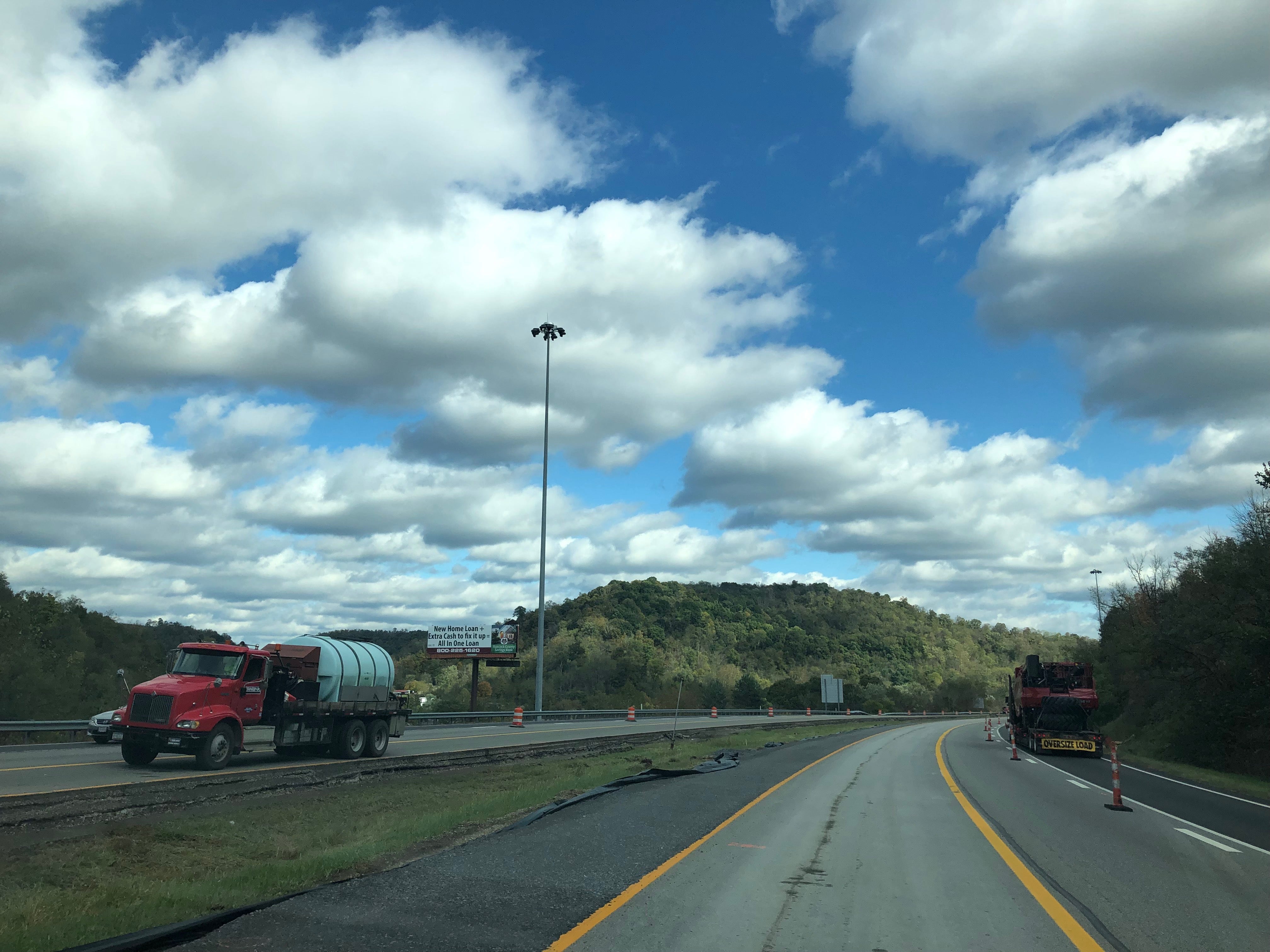 A red water truck traveling through the I-70 construction corridor in Wheeling against a backdrop of rolling hills and a cloudy sky.