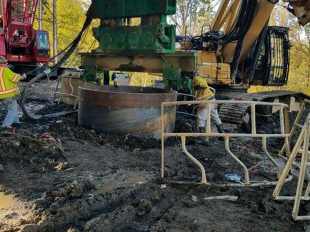 Construction crew using a green vibratory hammer to install a large steel foundation casing for the I-70 Fulton Bridge replacement in Wheeling.