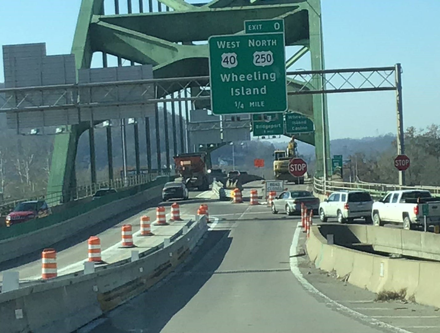 Westbound traffic on I-70 approaching Exit 0 (Wheeling Island) on the Fort Henry Bridge in Wheeling, WV, during the I-70 Forward bridge rehabilitation project. The image shows narrowed lanes marked by orange construction barrels and a green overhead exit sign for US 40 and US 250.