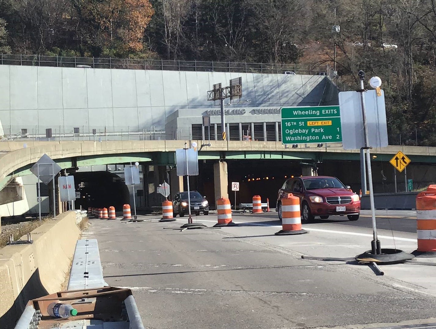 View of the Eastbound I-70 approach to the Wheeling Tunnel during the I-70 Forward project. A green "Wheeling Exits" sign lists 16th Street, Oglebay Park, and Washington Avenue, surrounded by orange traffic barrels.