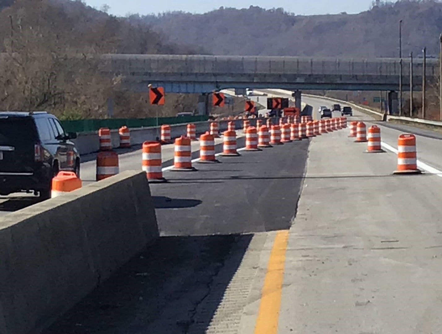 A long line of orange and white construction barrels with chevron signs tapering traffic on Interstate 70 in Wheeling during the initial phases of the I-70 Forward bridge rehabilitation project. An overpass is visible in the background against a hilly landscape.