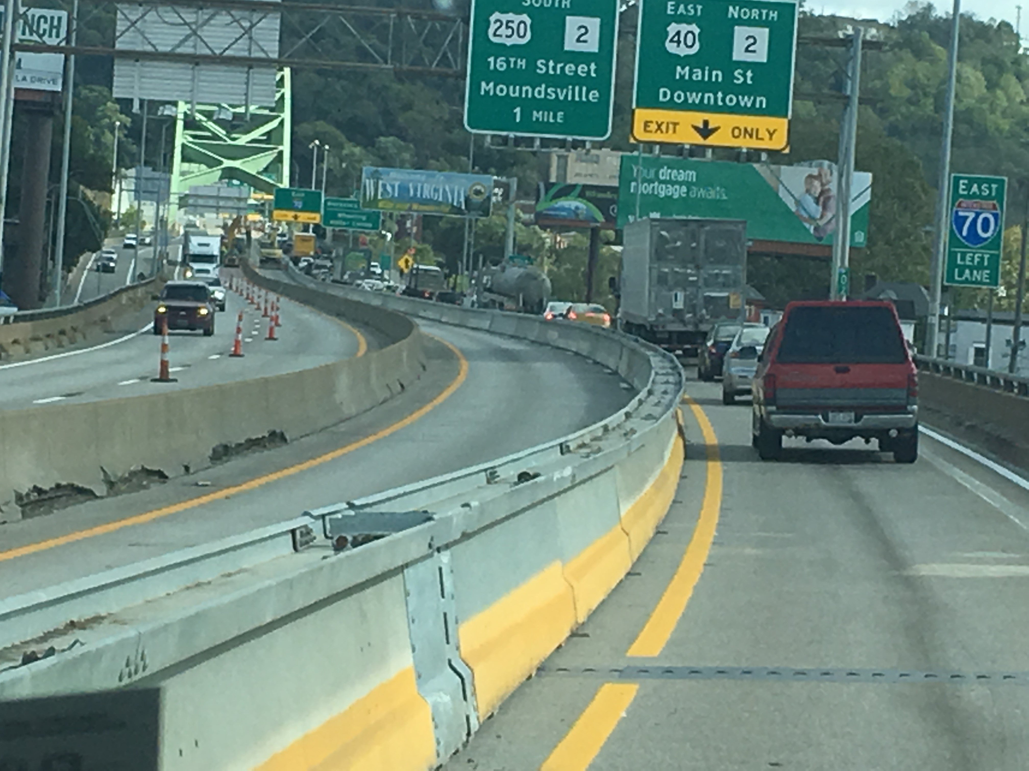 View of traffic on I-70 Eastbound in Wheeling, WV, showing highway exit signs and concrete barriers within the bridge construction zone.