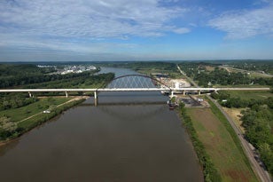 A high-angle aerial view of the modern Blennerhassett Island Bridge, featuring its distinctive rust-colored steel tied-arch main span and long concrete approach viaducts crossing the wide Ohio River.