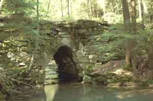 A view of the historic Clendenin Stone Arch Bridge built from rough-hewn stones, spanning a small creek in a dense, sun-dappled forest.