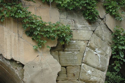 A close-up detail of the historic Elm Grove Stone Arch Bridge, showing cracked concrete parging peeling away to expose the underlying stone blocks and overgrown green ivy vines.