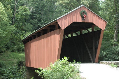 A three-quarter view of the historic Fletcher Covered Bridge, featuring its red vertical wooden siding and a holiday wreath hanging above the open portal entrance, surrounded by thick green foliage.