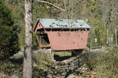 A side view of the red wooden Laurel Creek Covered Bridge spanning a rocky creek bed surrounded by trees