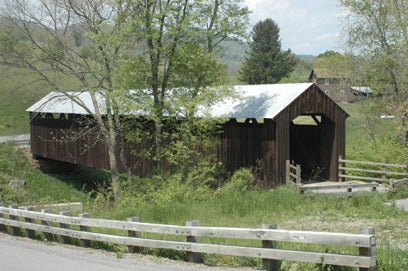 Side view of the rustic, dark wooden Locust Creek Covered Bridge surrounded by trees in a rural setting