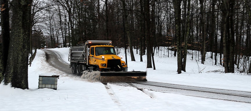 A yellow snowplow truck clearing slush and snow from a rural tree-lined road