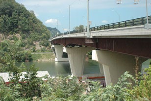 A three-quarter low-angle view of the modern Star City Bridge, highlighting its dark steel plate girders and flared concrete piers spanning a wide river with forested hills in the background.