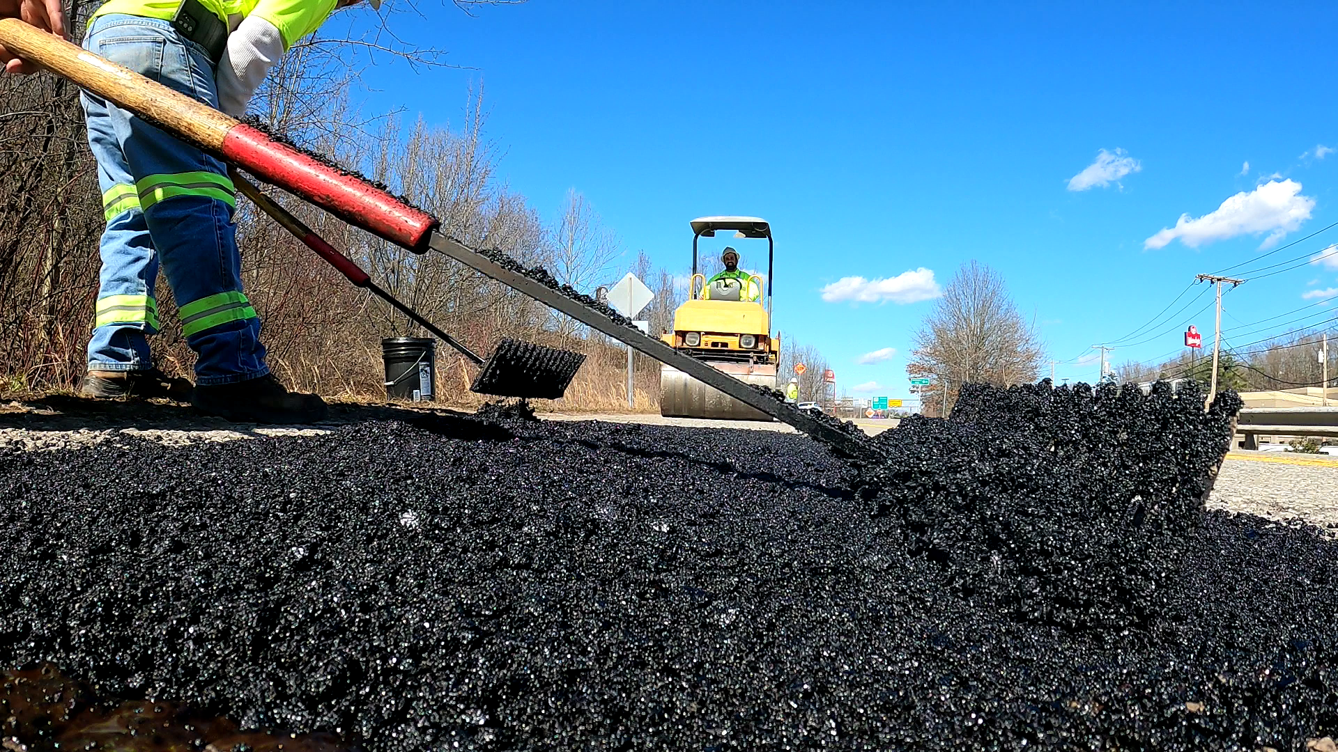 A road crew worker in high-visibility gear uses a rake to spread fresh black asphalt while another worker operates a yellow steam roller in the background