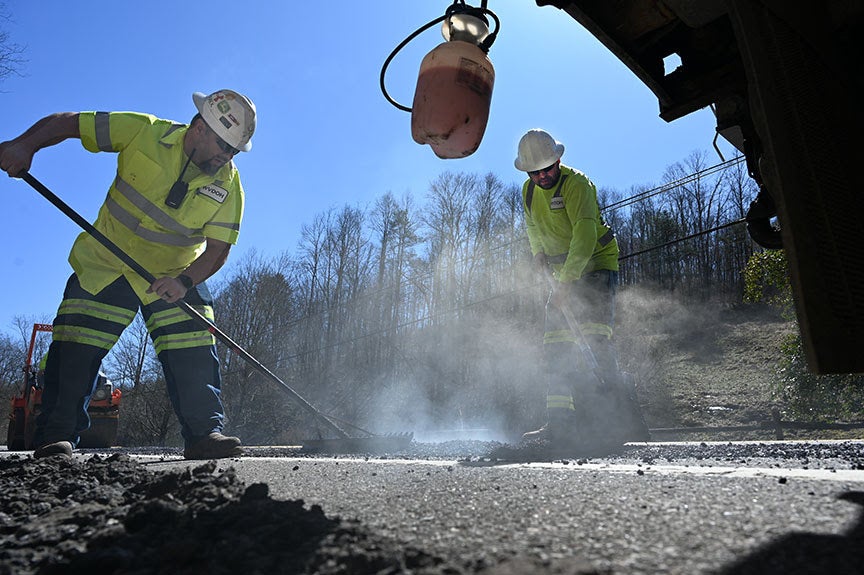 Two WVDOH road crew workers in hard hats and high-visibility shirts rake and shovel steaming hot asphalt to patch a roadway.