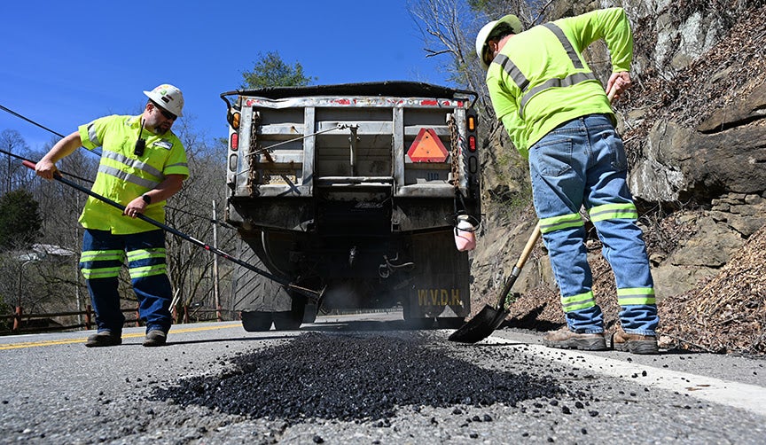 Two WVDOH road workers in high-visibility gear use a rake and shovel to fill a road trench with fresh asphalt behind a parked dump truck.