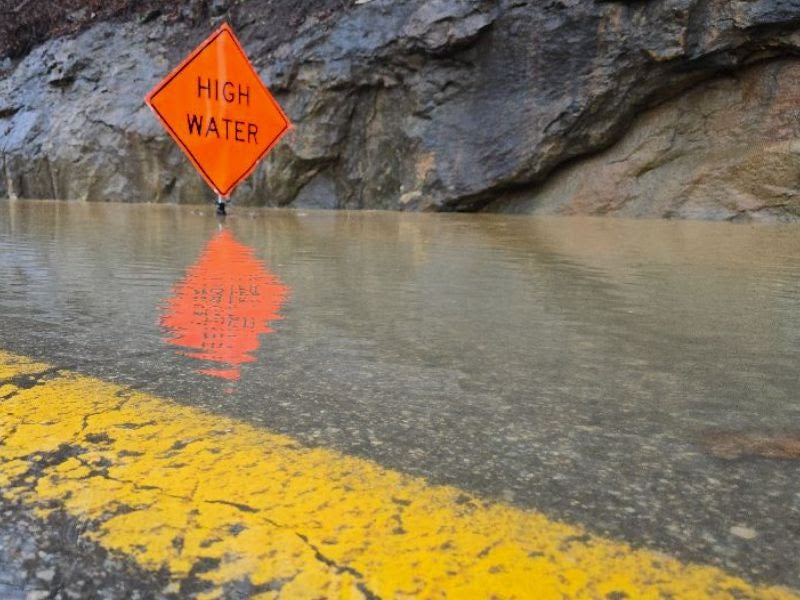 An orange diamond warning sign reading 'High Water' stands on a completely flooded road next to a rocky cliffside, reflecting in the murky floodwater