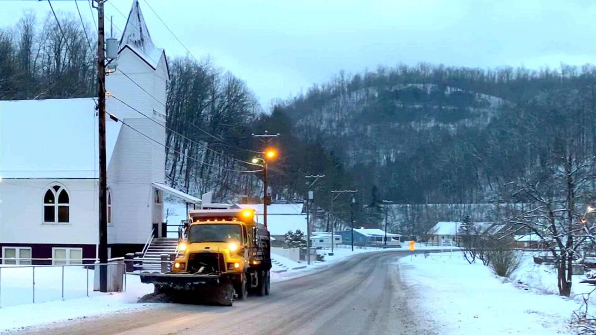 A yellow WVDOH snowplow truck with amber lights flashing on a snow-covered rural road next to a white country church