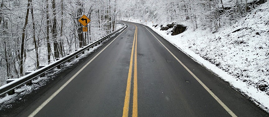 A wet, cleared two-lane road curving through snow-covered trees with a yellow 15 mph curve warning sign