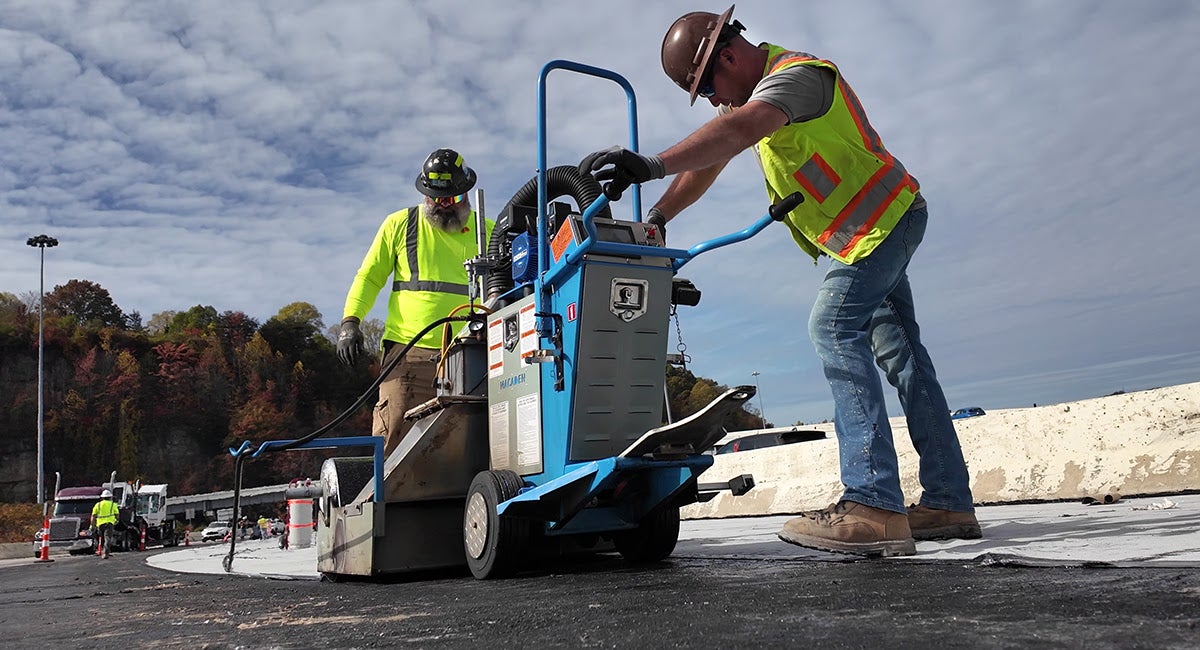 Two construction workers operate an automated machine to apply a protective waterproofing membrane layer to the bridge deck.