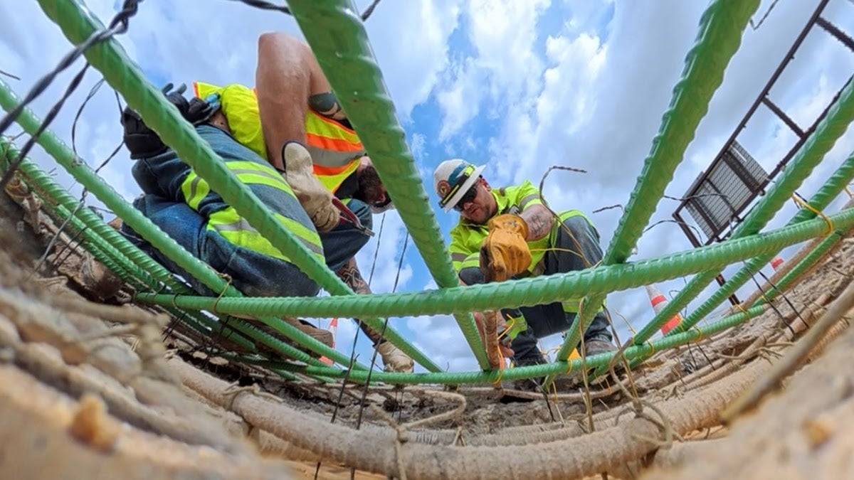 A low-angle view looking up from inside a large pothole at two highway workers in high-visibility gear securing a grid of green rebar against a partly cloudy blue sky.