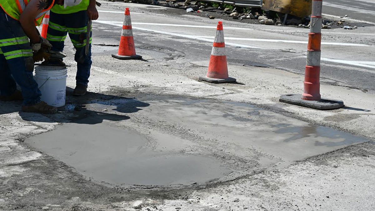 A large rectangular patch of freshly poured, wet concrete filling a pothole on a roadway, cordoned off by orange traffic cones and delineator posts.
