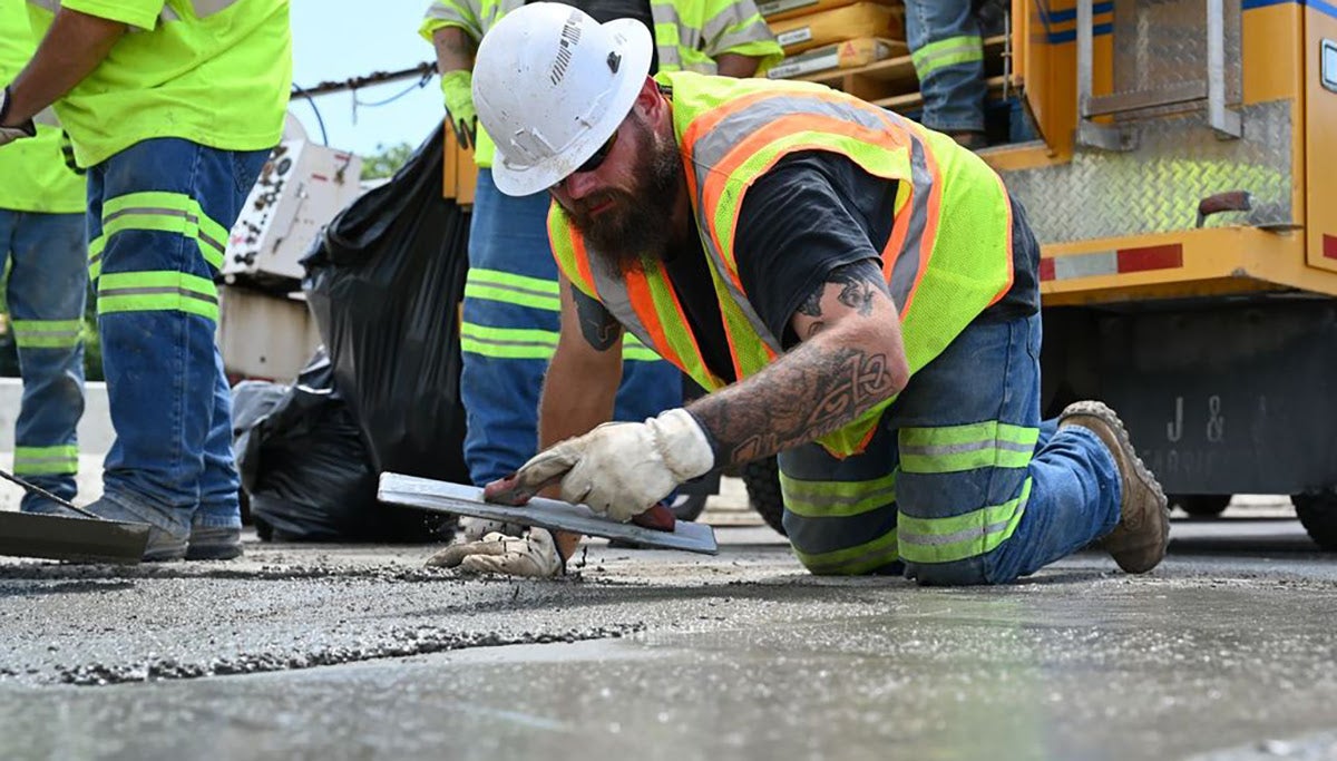 A highway construction worker kneeling on the road uses a hand trowel to smooth out wet concrete over a newly patched pothole.