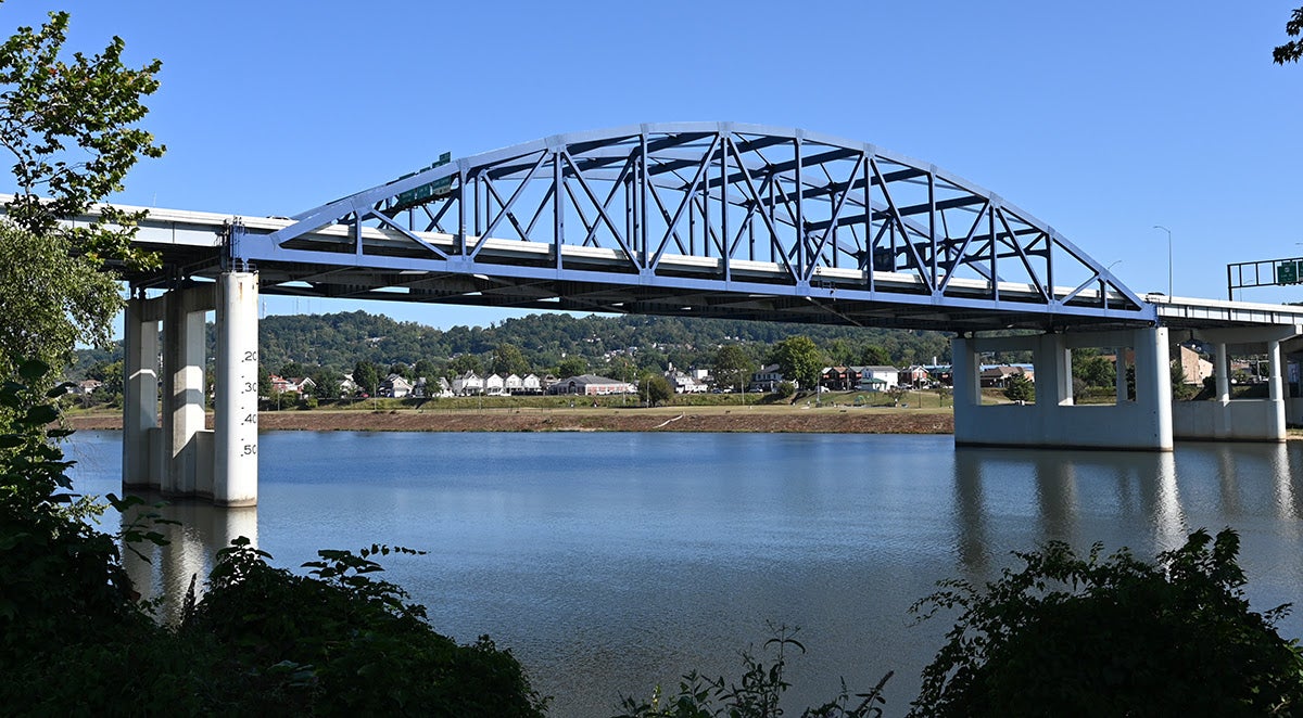 Blue steel arch truss bridge spanning a wide river with hills in the background.