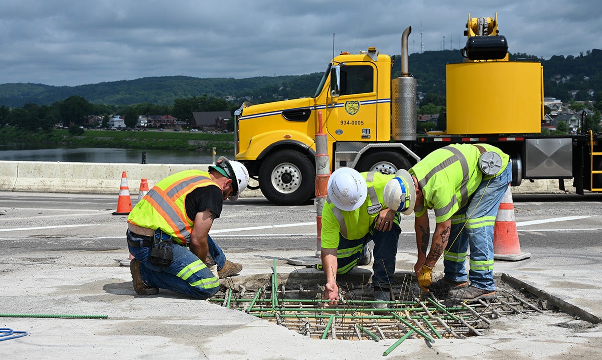 Three construction workers in safety vests repair reinforcement rebar on a bridge deck in front of a yellow utility truck.