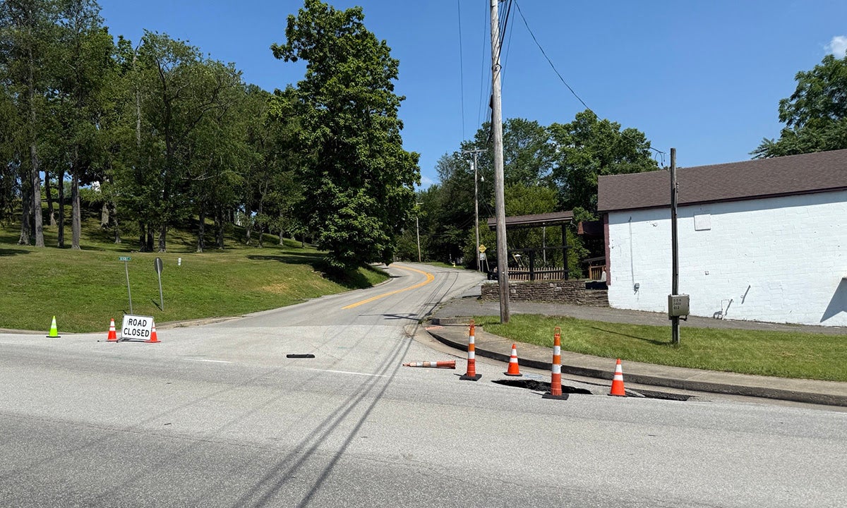 A sinkhole forming at a paved road intersection, cordoned off by orange traffic cones next to a "Road Closed" barricade.