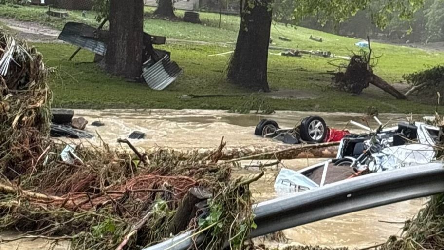 Fast-moving, muddy floodwaters filled with heavy debris, including uprooted trees and destroyed vehicles tangled against a mangled guardrail.