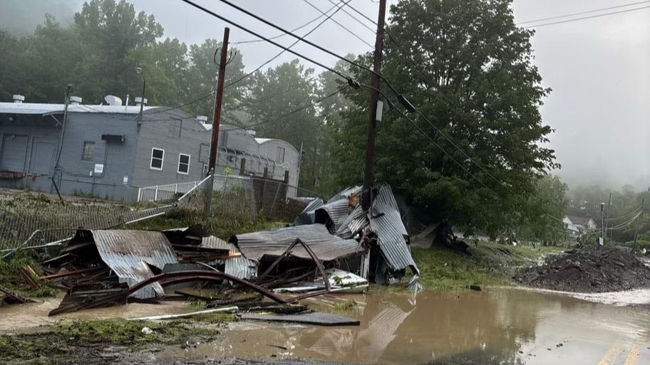 Muddy floodwaters covering a road next to the mangled corrugated metal remains of a destroyed building and a leaning utility pole.