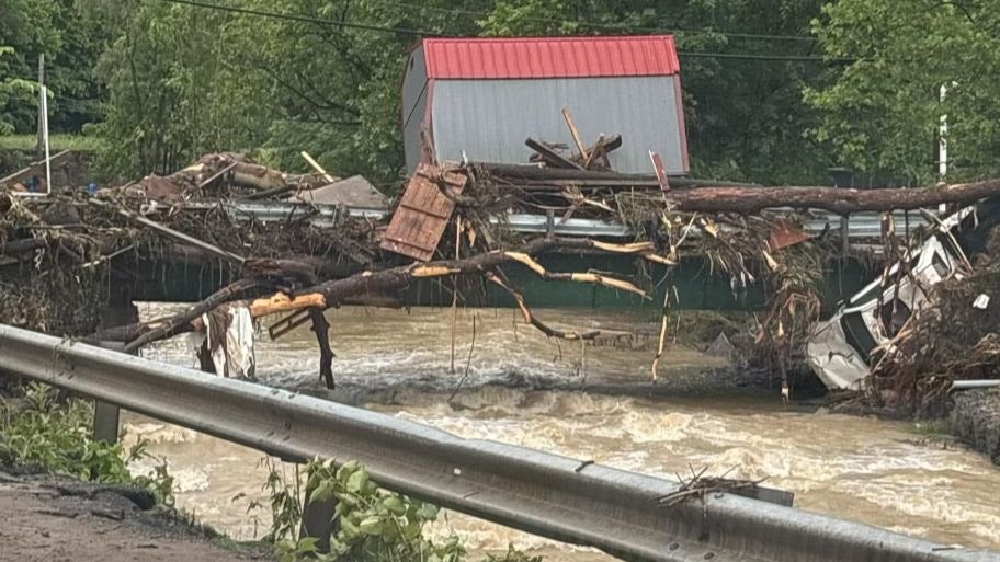 Massive flood debris, including uprooted trees, a small shed, and a white vehicle, completely blocking a bridge over fast-flowing, muddy water.
