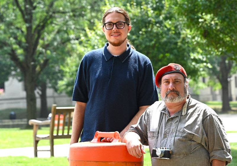 Two men posing outdoors next to an orange traffic barrel, with one wearing glasses and a blue polo shirt and the other wearing a red beret with a camera around his neck.