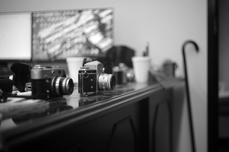 A black and white photograph showing several vintage film cameras sitting on a desk with a shallow depth of field, featuring a blurred computer monitor and a cane in the background.