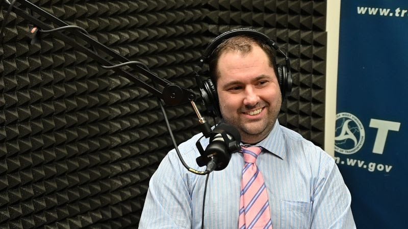 A smiling man in a light blue striped shirt and pink tie wearing headphones, positioned in front of a studio microphone inside a recording booth with acoustic foam wall panels.