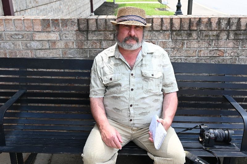 A man with a graying beard wearing a straw fedora hat and a short-sleeved button-down shirt, sitting on a black metal bench while holding a notepad, with a digital camera resting beside him.