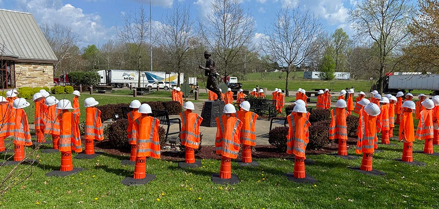 Dozens of orange WVDOH safety vests and white hard hats placed on traffic cones surround a bronze statue of a highway worker at the West Virginia Fallen Workers Memorial.