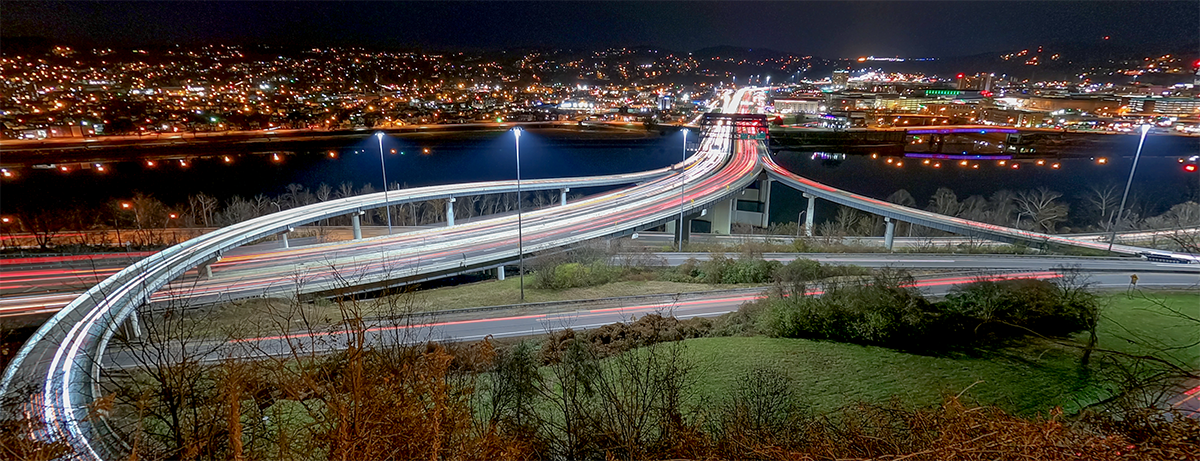 Panoramic night view of the Fort Hill bridge and highway interchange in West Virginia, featuring long-exposure white and red light trails from traffic sweeping across curving elevated ramps over a river, with a brightly lit city skyline and hillsides in the background.