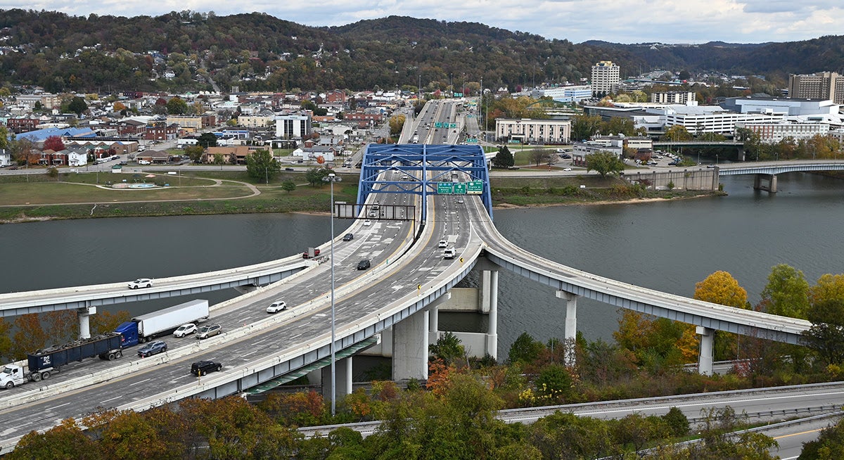 Elevated daytime view of the Fort Hill bridge and highway interchange, featuring a prominent blue steel truss bridge spanning a wide river. Cars and semi-trucks travel along curving concrete ramps that lead into a city skyline backed by rolling, tree-covered hills under a cloudy sky.