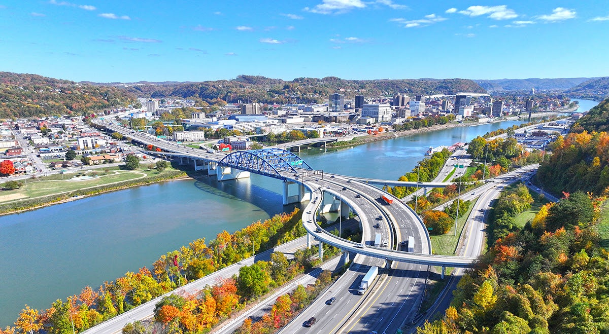 Wide aerial view of the Fort Hill bridge and highway interchange crossing a wide river. Curving concrete ramps carry traffic through hillsides covered in vibrant autumn foliage, leading toward a downtown city skyline under a clear blue sky.