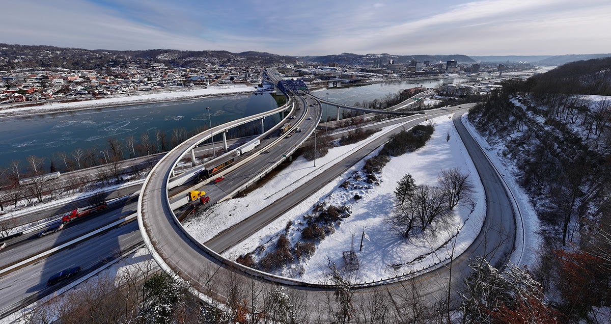 High-angle winter view of the Fort Hill bridge and highway interchange in Charleston, West Virginia, with snow covering the surrounding hillsides and riverbanks. Semitrucks and cars travel along a large, sweeping concrete ramp leading toward a blue steel truss bridge over the river, with the downtown city skyline visible in the distant background.