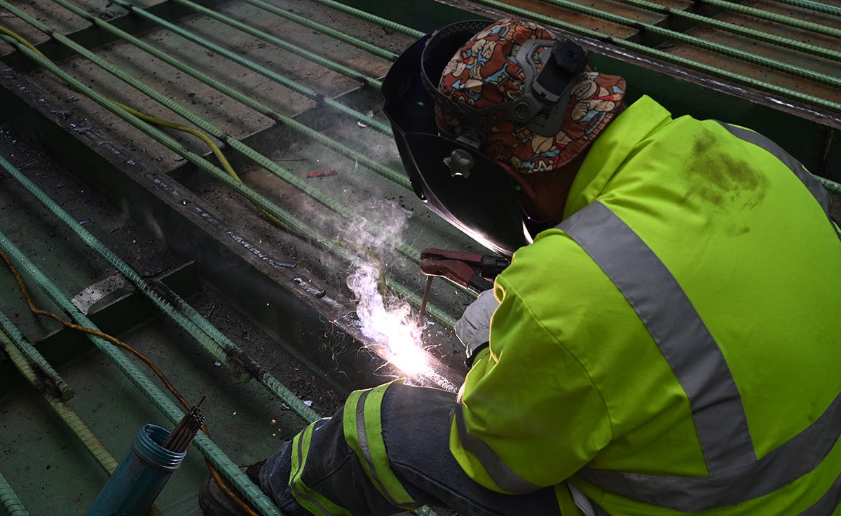 A welder wearing a protective mask and high-visibility jacket welds the steel grid framework of a bridge deck.