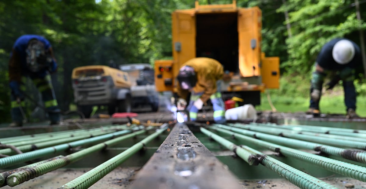 A low-angle view focuses on the steel rebar framework as construction workers weld in the background.
