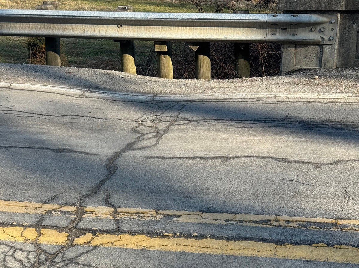 A close-up view of heavily cracked and dipping asphalt pavement on a two-lane road running alongside a metal guardrail